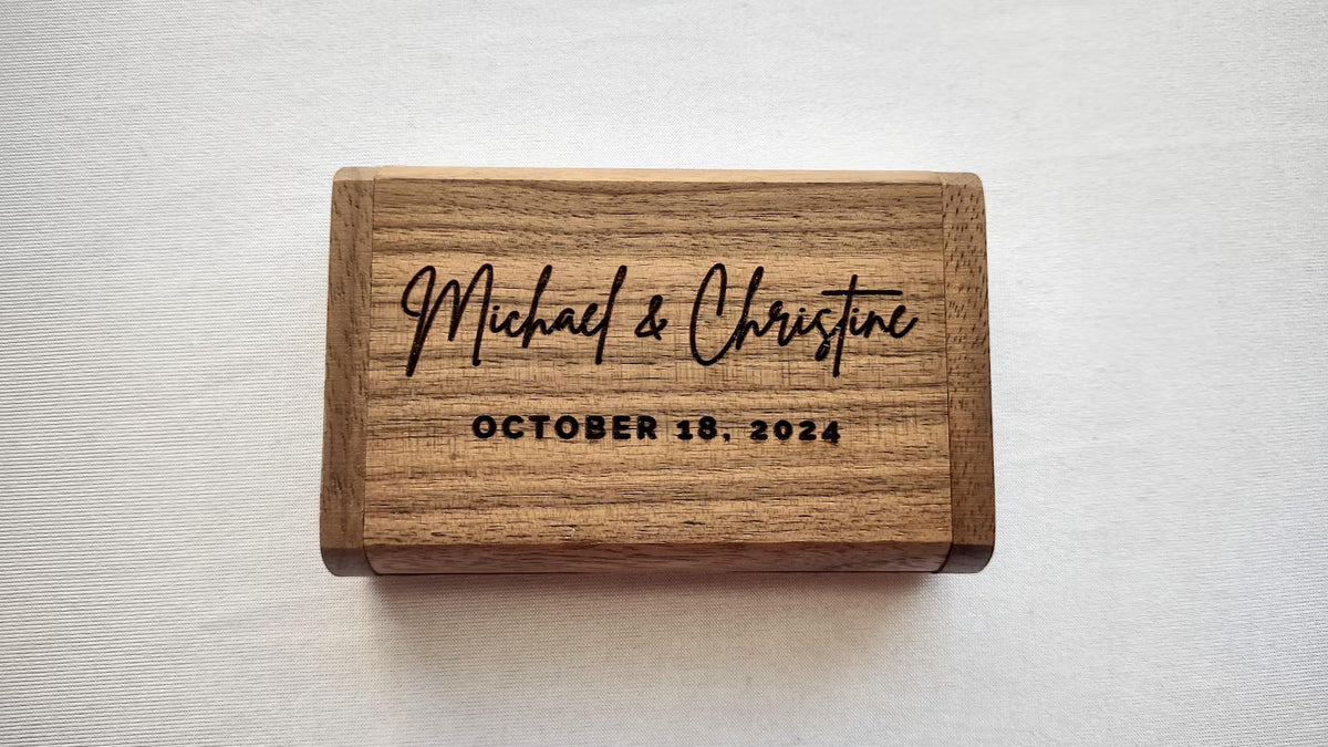A rectangular wooden keepsake box with engraved black script reading 'Michael & Christine' and 'OCTOBER 18, 2024', photographed from above on a light neutral background.